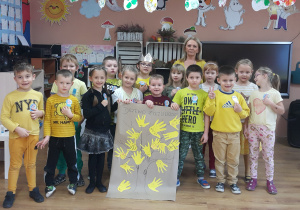 Grupa dzieci wraz z nauczycielem ubranych na żółto, wspólnie prezentują ilustrację z wykonanym "Drzewem Życzliwości"."
