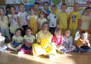 Dzieci ubrane na żółto, stoją lub siedzą, wśród nich siedzi nauczycielka z żółtym balonem.