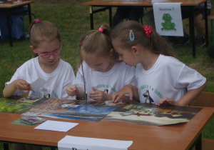 Trzy dziewczynki w białych koszulkach oglądają ilustracje.