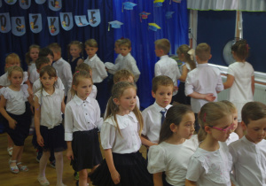 Dzieci w białych bluzkach tańczą na tle granatowej dekoracji.